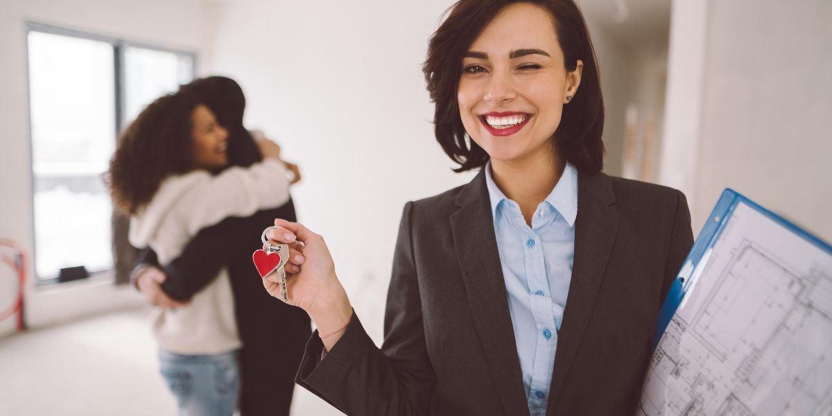 Smiling real estate agent holding house keys with a couple hugging in the background.