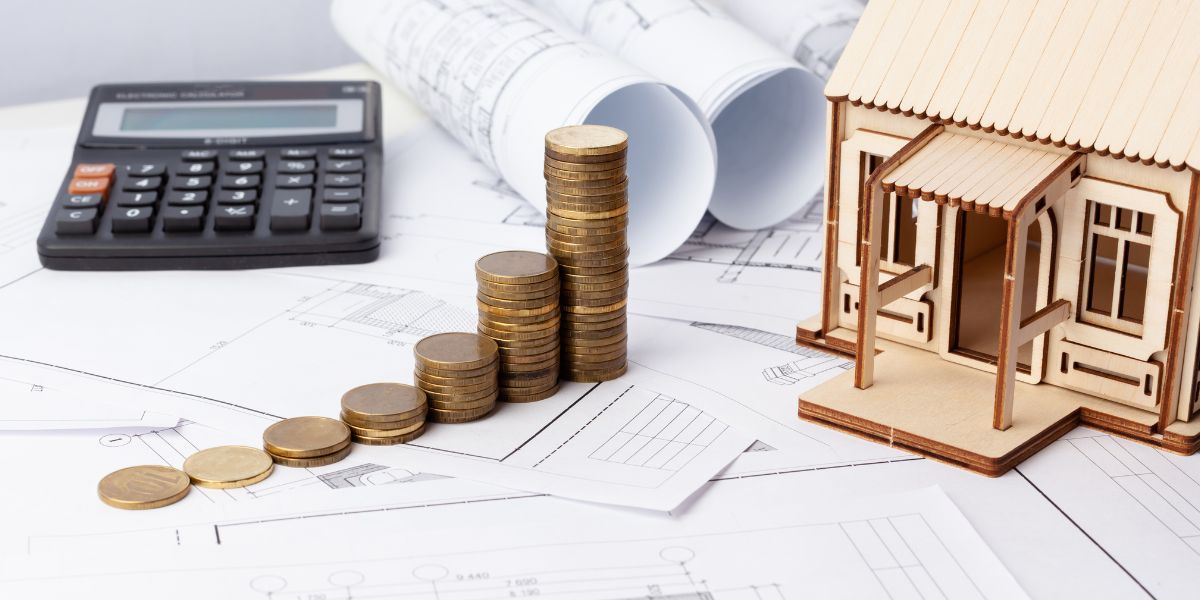 Stacks of coins, a calculator, and a small wooden house model on architectural blueprints.